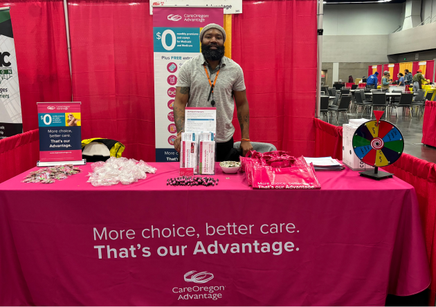 Promotional Event Booth for CareOregon Advantage, adorned with promotional items, a prize wheel and manned by an individual standing behind the table, wearing a lanyard